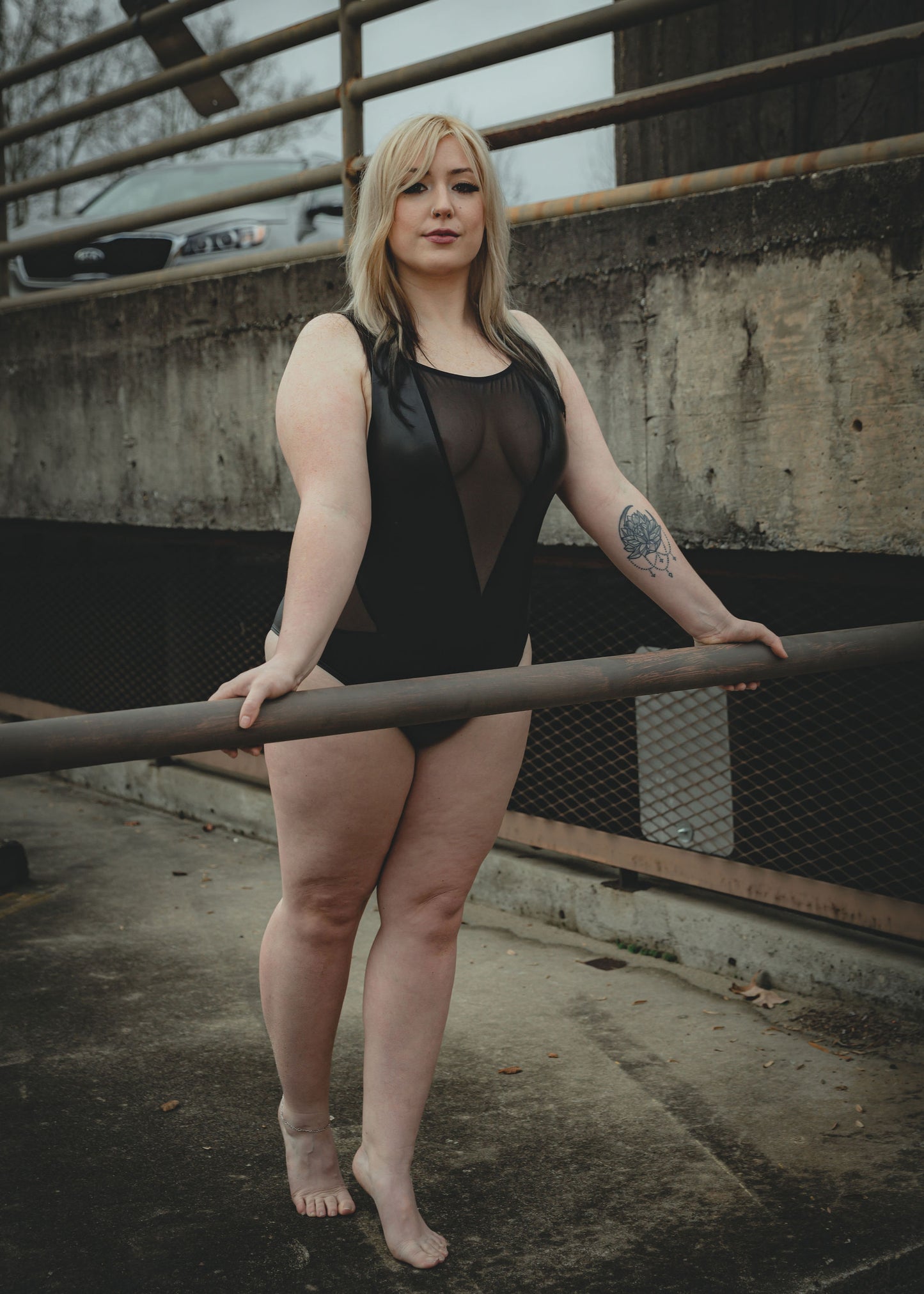 Image of high quality black faux leather ethical bodysuit with mesh accents on a woman who is posed against railing.