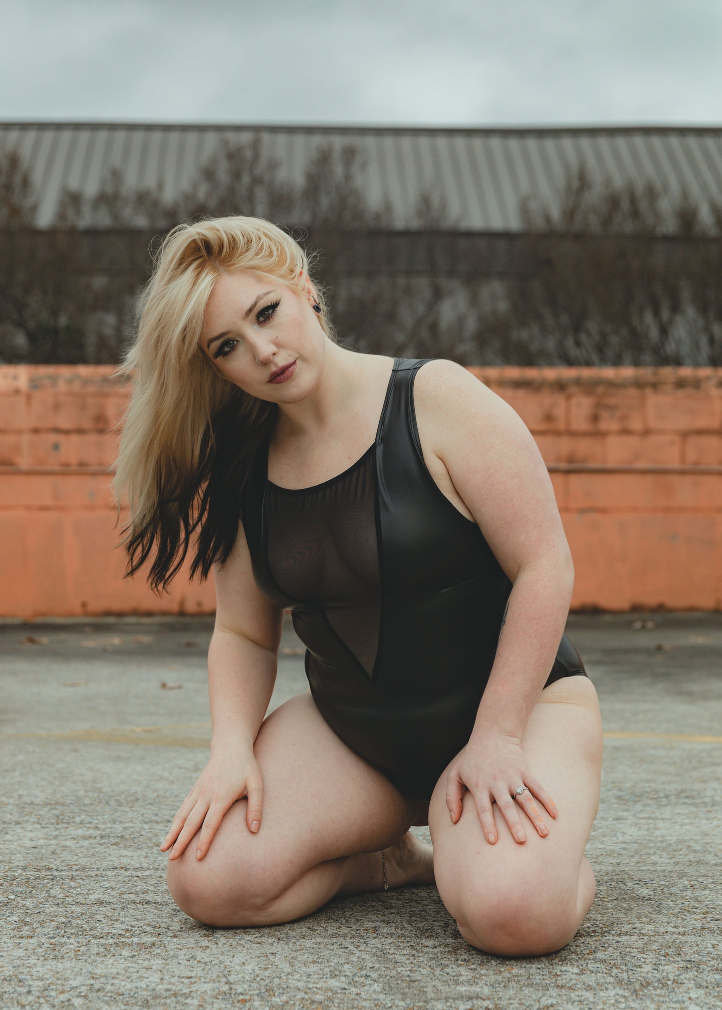 Image of high quality ethical black faux leather bodysuit with mesh accents on a woman who is knelt down on asphalt, posed.
