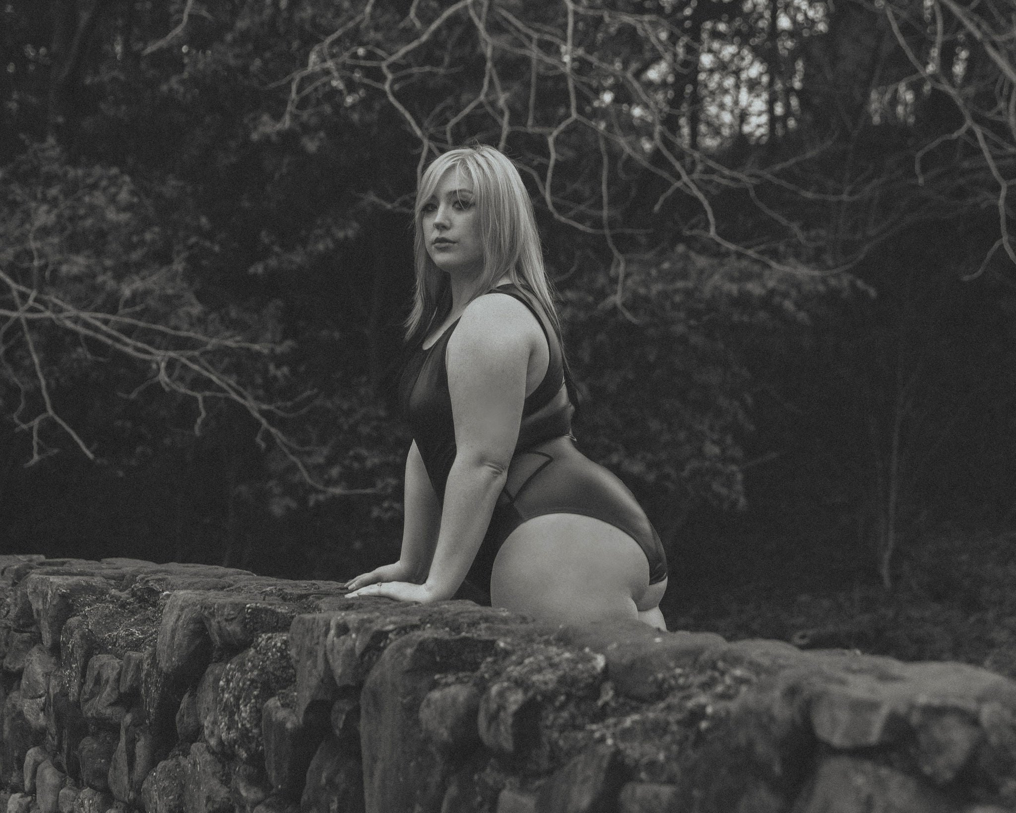 Black and white image of woman standing outdoors by a rock wall wearing a leather bodysuit.